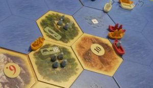 A board game of Settlers of Catan is shown with land and and sea tiles. At the edges of the sea files there are ships transporting people and spices!